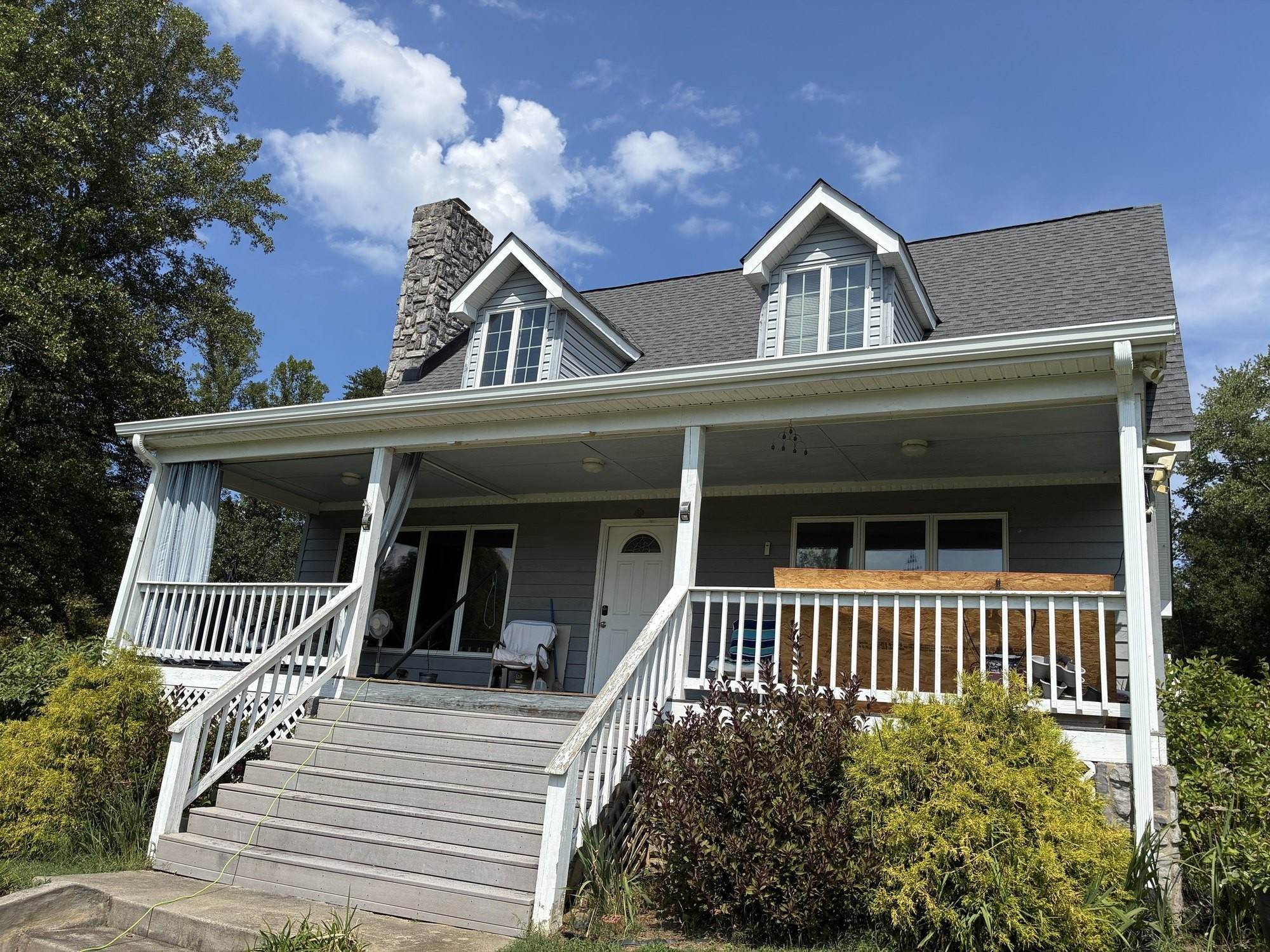 Gray Cape Cod home with two dormers, a stone chimney and a wide front porch under a new charcoal architectural shingle roof