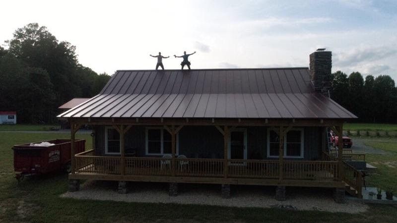 Two roofers standing on the ridge of a finished dark metal roof over a wraparound porch